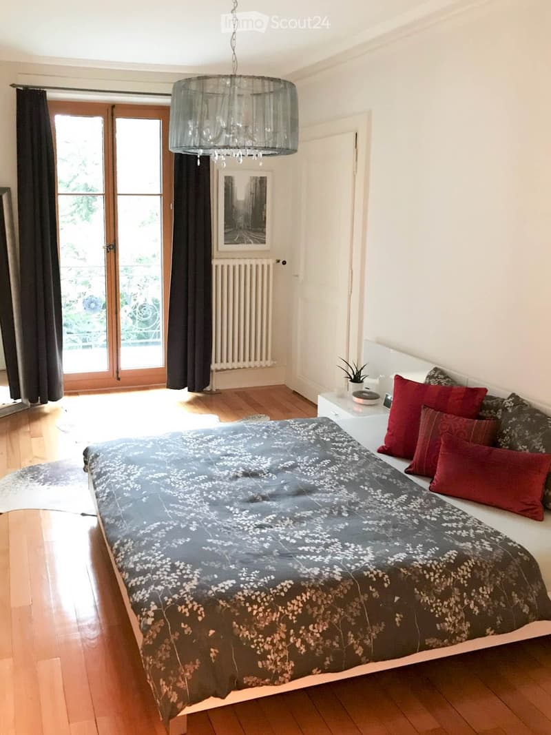 Bright bedroom with wooden flooring, large bed with grey and white patterned duvet and red pillows, nightstand with plant, chandelier, large window with dark curtains, radiator, and picture frame on wall.