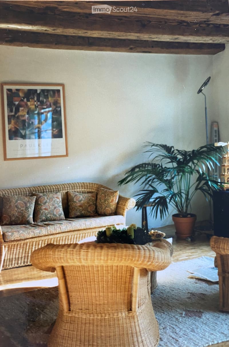 A cozy living room with a wicker sofa, patterned cushions, a wicker coffee table with a decorative centerpiece, a wicker armchair, a tall potted plant, and a picture frame on the wall.