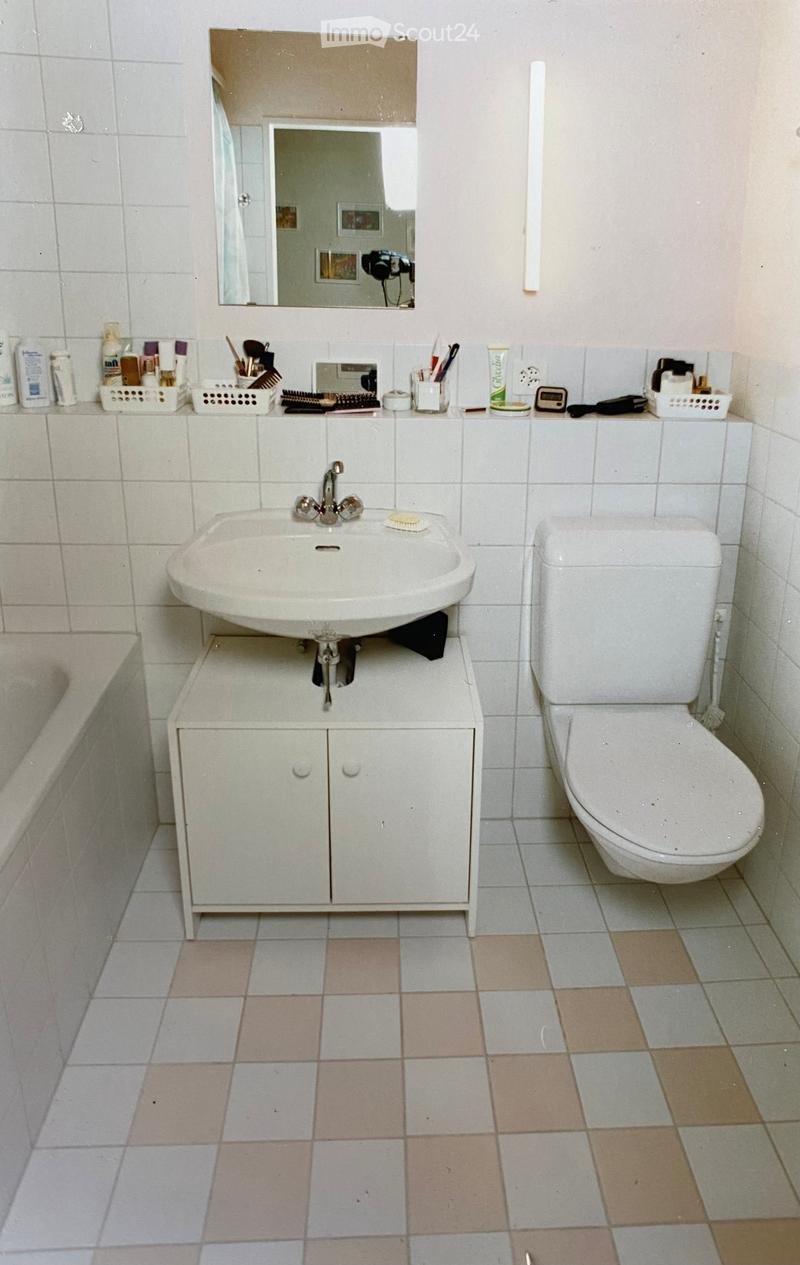 A small bathroom with a white sink, toilet, and mirror. The sink is mounted on a white cabinet with two doors. The floor is tiled with a mix of light and dark tiles. Various toiletries and personal care items are placed on the sink counter, including a to