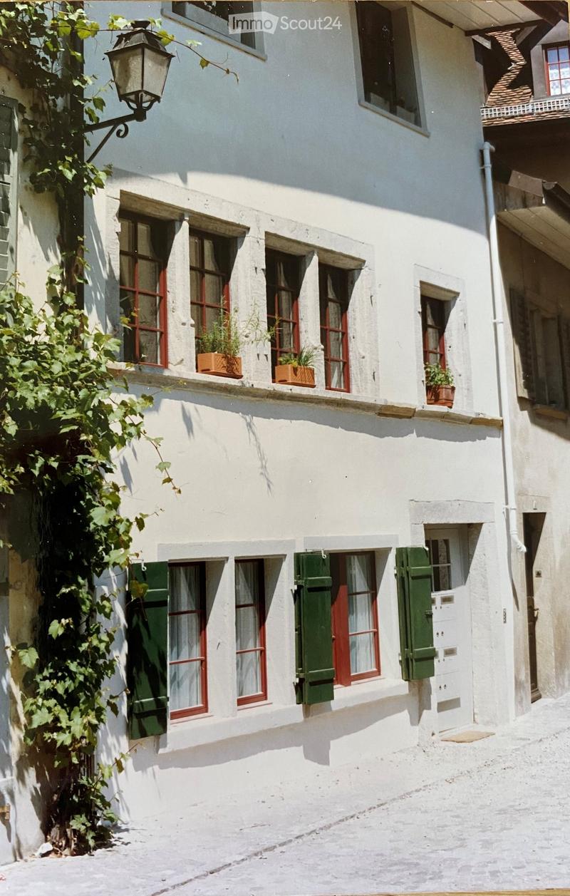 Two-story white building with red-framed windows, green shutters, and potted plants on the windowsill.