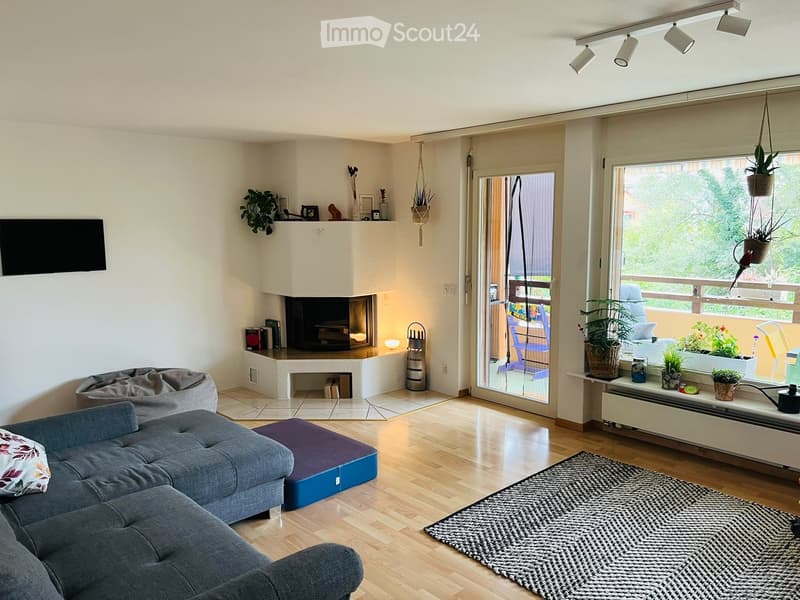 Modern living room with a grey sectional sofa, ottoman, fireplace, large windows, and a balcony with outdoor furniture.