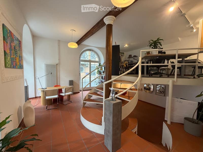 Spacious, open-plan living area with terracotta tiles, a curved staircase, a dining table with chairs, a decorative vase, and a potted plant. The room features exposed wooden beams and large arched windows.