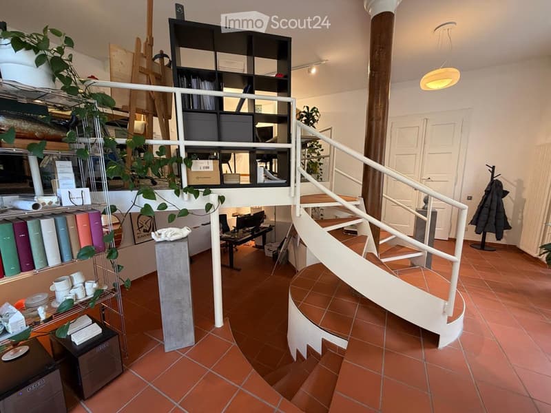 A modern, open-concept room with terracotta flooring, a spiral staircase leading to an upper level, shelves filled with books and decorative items, and a small seating area with a chair and table.