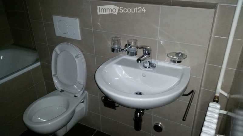Bathroom with a toilet, sink, and faucet. The sink has a modern design with a chrome faucet and two small cups placed on the countertop. The walls are tiled in a neutral color, and a small shelf is visible above the sink.