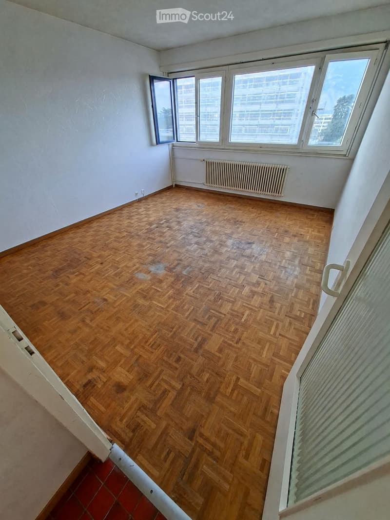Empty room with wooden herringbone flooring, white walls, large windows, and a radiator. No furniture or equipment present.