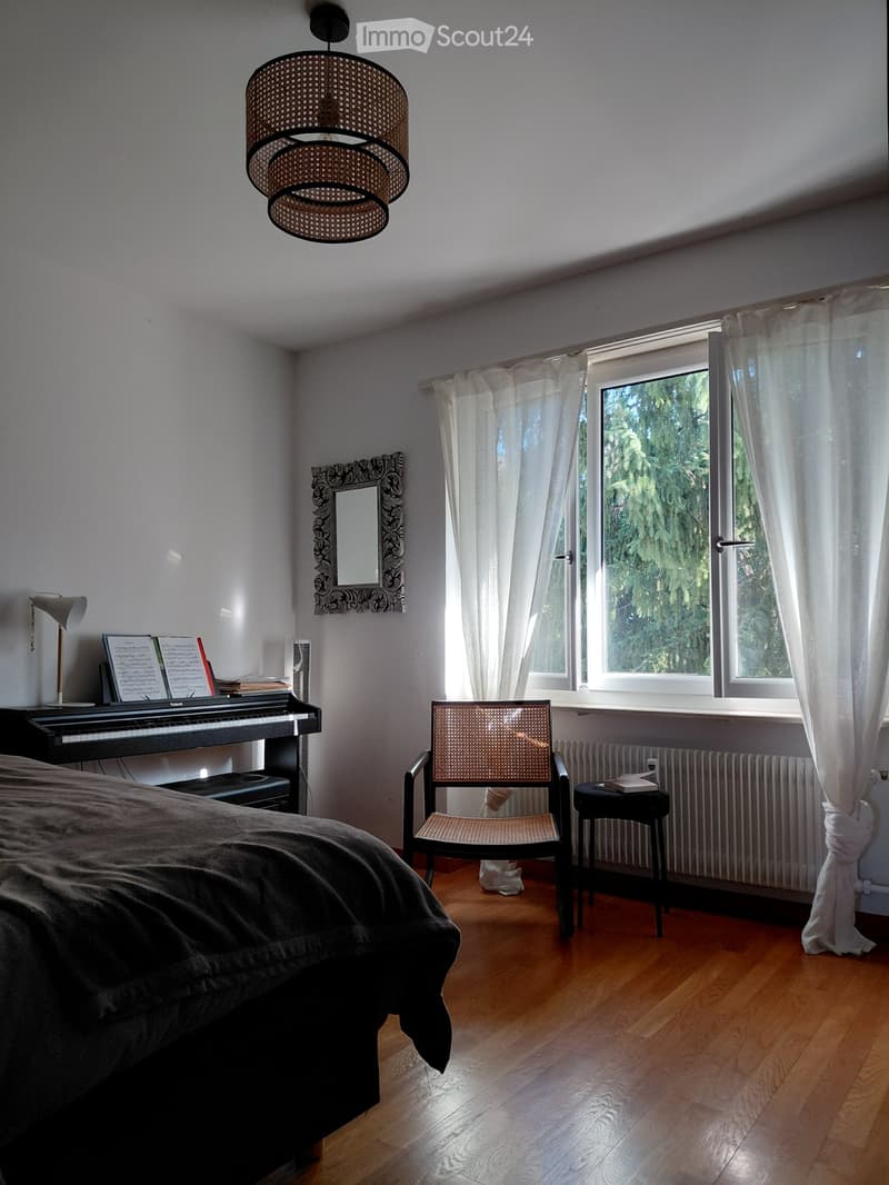 Cozy bedroom with wooden flooring, large window with white curtains, bed with dark bedding, wicker chair, small black stool, piano, mirror, and hanging woven lamp.