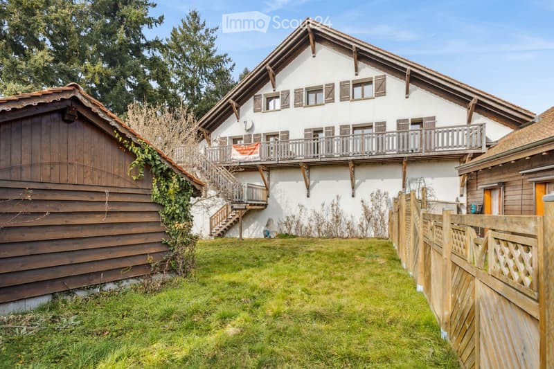 Two-story house with white exterior, wooden balcony, and wooden garage. The house features multiple windows with shutters and a well-maintained lawn in front.