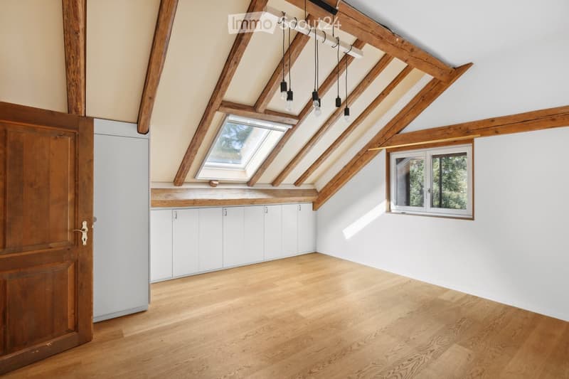 Bright, empty attic room with wooden beams, skylight, and white cabinetry. Wooden floor and white walls.