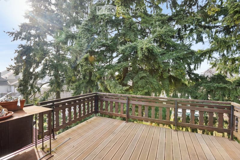 Wooden balcony with railing, overlooking a lush green tree-filled area. The deck is made of light-colored wood planks, and the railing features vertical wooden balusters.