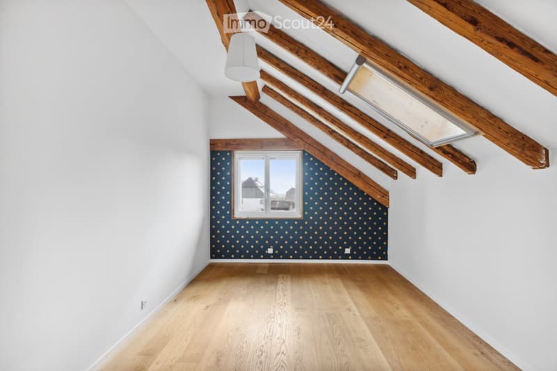 Empty attic room with wooden beams, wooden flooring, white walls, and a window with a view of the outside.