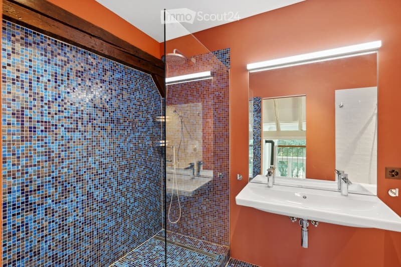 Modern bathroom with orange walls, a glass-enclosed shower with blue mosaic tiles, a double sink, and a large mirror.