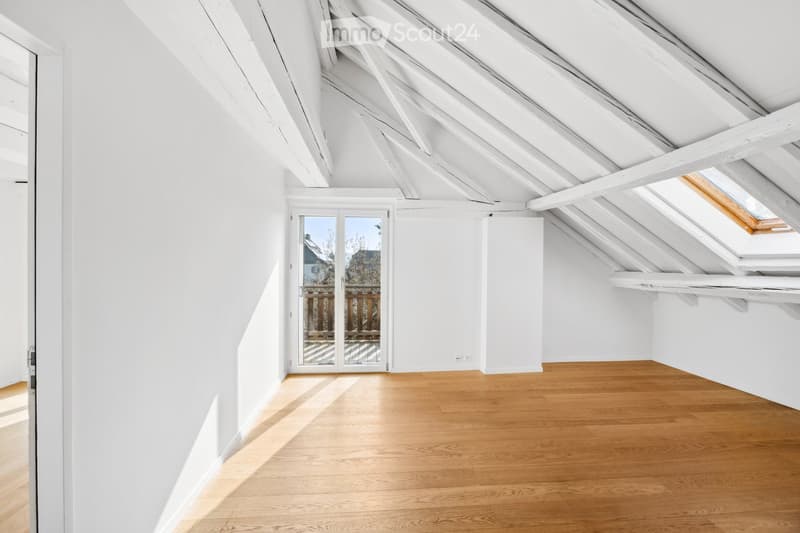 Bright, empty attic room with wooden flooring, white walls, and exposed wooden beams. Features a large skylight and a glass door leading to an outdoor deck.