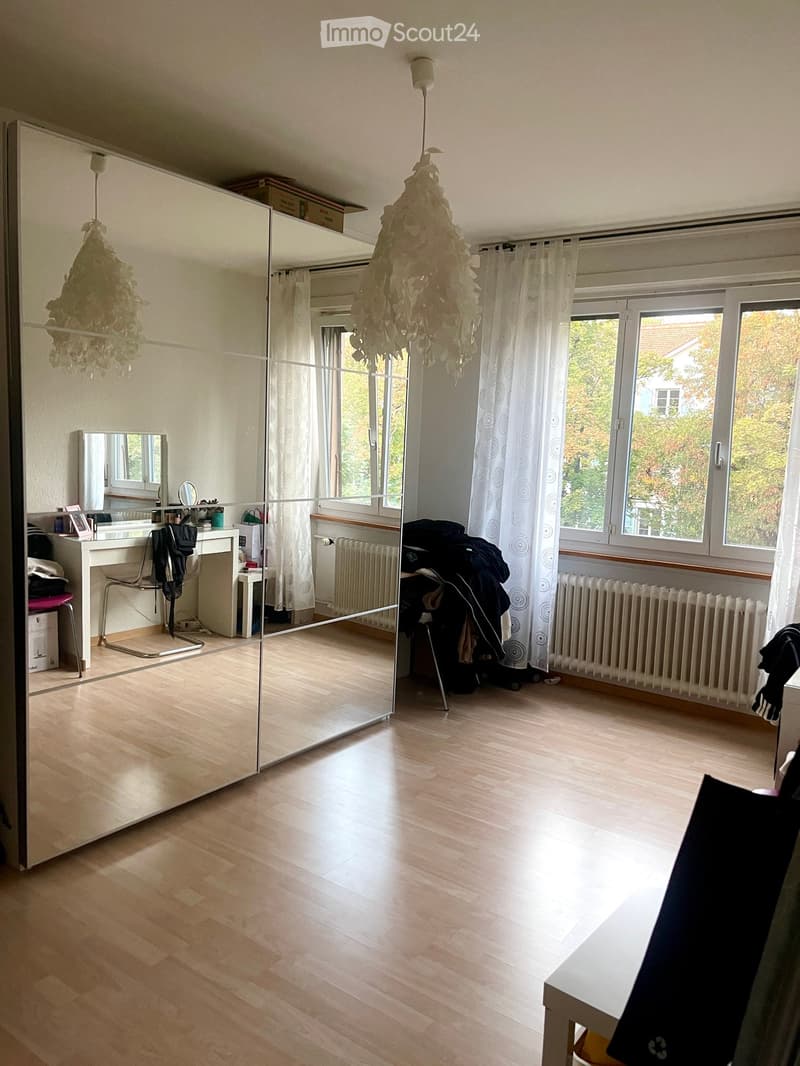 Well-lit room with wooden flooring, large mirror, and a small desk area with a chair and mirror. Natural light streams in through large windows with white curtains.
