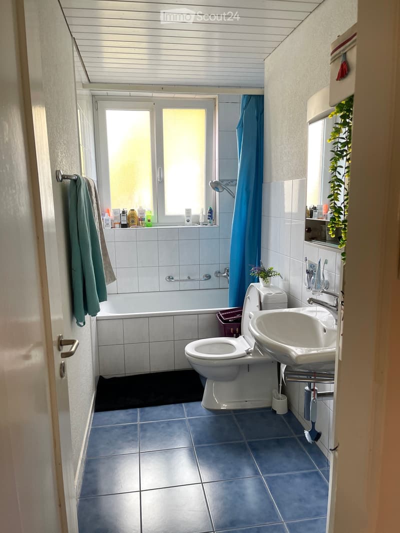 Bathroom with blue tiles, white fixtures, a bathtub, toilet, sink, and a window with a blue shower curtain.
