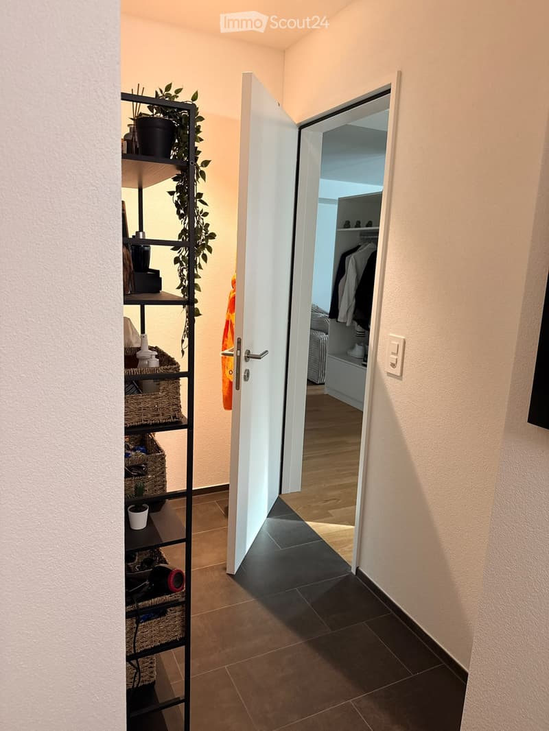A modern hallway with beige walls, dark tiled floor, and an open door leading to a room with wooden flooring and a closet with hanging clothes.