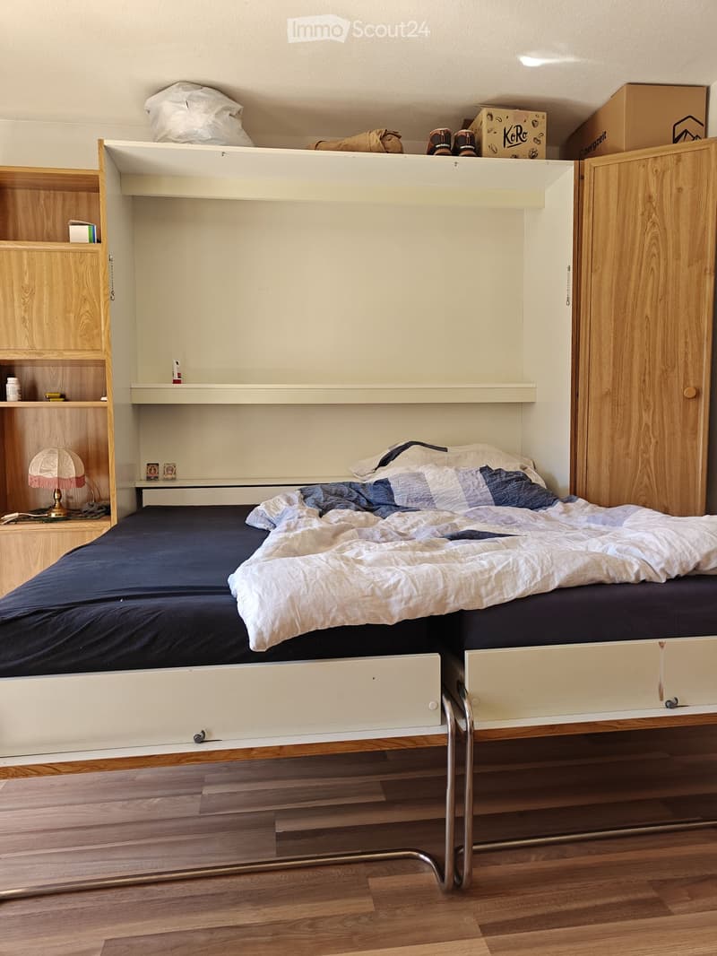 A compact bedroom with a lofted bed, wooden floor, built-in shelves, and a small nightstand with a lamp.