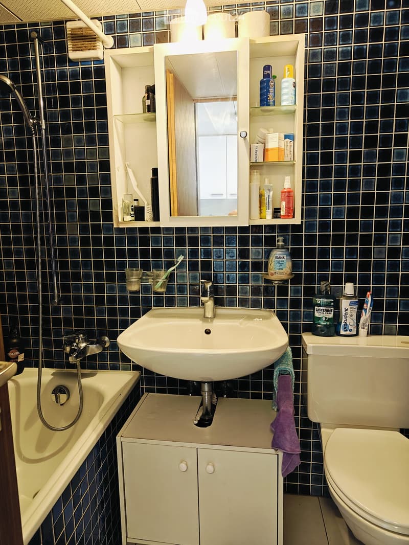 Bathroom with dark blue tiled walls, a white sink, a toilet, a bathtub, a mirror with shelves, and various toiletries.