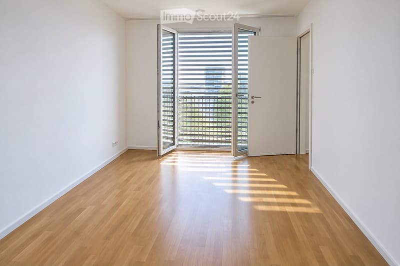 Empty room with light wooden flooring and white walls, featuring an open sliding door leading to a balcony with a view of greenery.