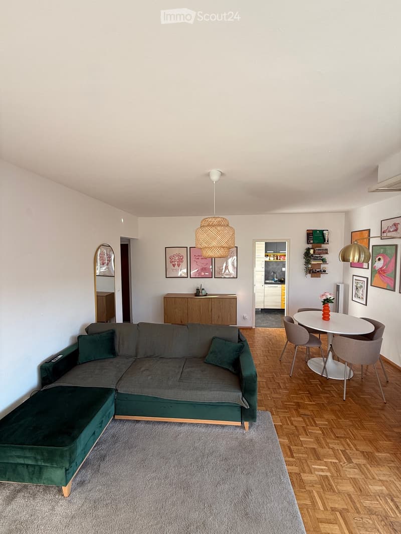 A modern living room with a grey sectional sofa, a round dining table with four chairs, a wooden cabinet, a hanging lamp, and several picture frames on the wall.