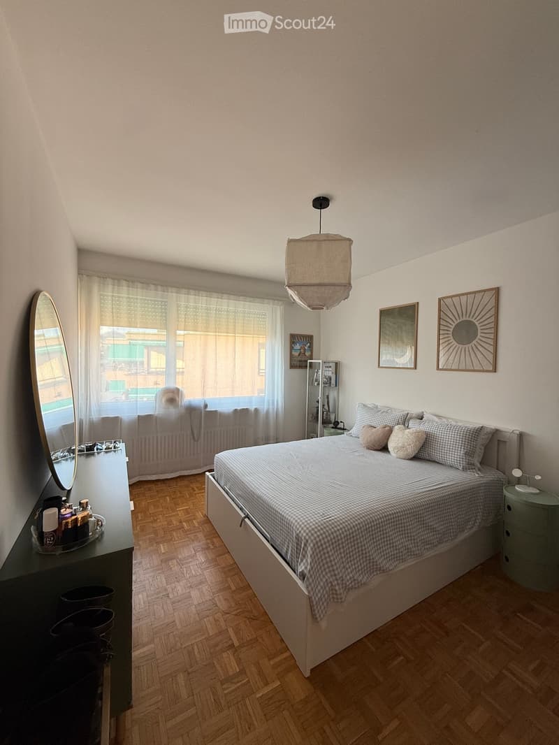 A modern bedroom with a wooden floor, a large bed with checkered bedding, a nightstand with a lamp, a mirror, and several picture frames on the wall. A window with curtains offers a view of the street.