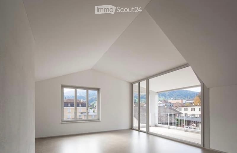 Bright, empty room with white walls and a sloped ceiling, featuring large windows and a sliding glass door leading to a balcony with a scenic view.