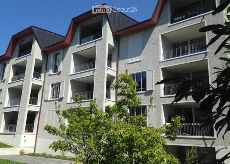 Modern multi-story apartment building with grey facade, white shutters, and multiple balconies. The building features a red roof and is surrounded by greenery.