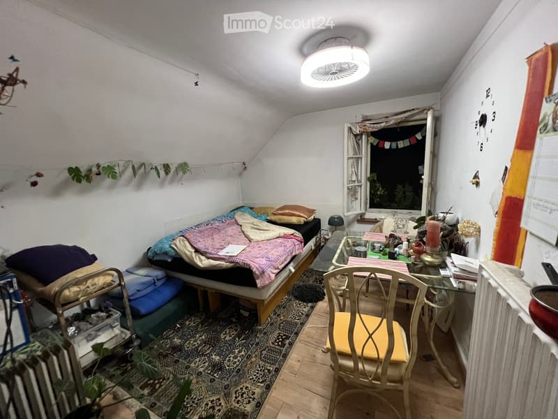 Cozy bedroom with a slanted ceiling, featuring a bed with colorful blankets, a small table with a lamp, a chair, and a window with a view of greenery.