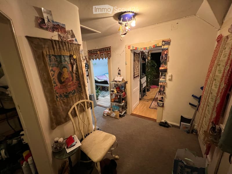 A small, lived-in room with a chair, small table, shelf, and various decorations including a large tapestry and a mirror. The room has carpeted flooring and a ceiling light fixture.