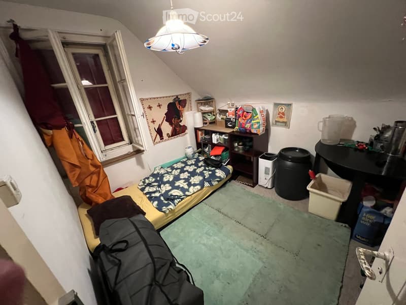 Small attic bedroom with a single bed, small table, chair, shelf, and various personal items. The room has slanted ceilings and a window with an open curtain.