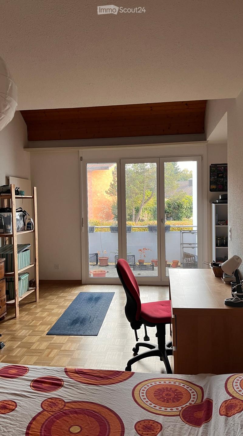 Brightly lit room with wooden flooring, a bed with a colorful quilt, a red office chair, a desk with a lamp, and shelves with various items. Large sliding glass doors lead to a balcony with potted plants.