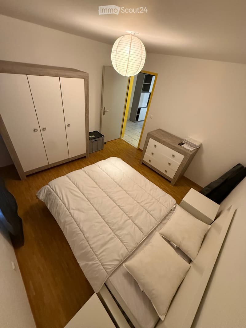 Modern bedroom with a neatly made bed, white bedding, a nightstand with drawers, a wardrobe, and a hanging paper lantern light fixture.