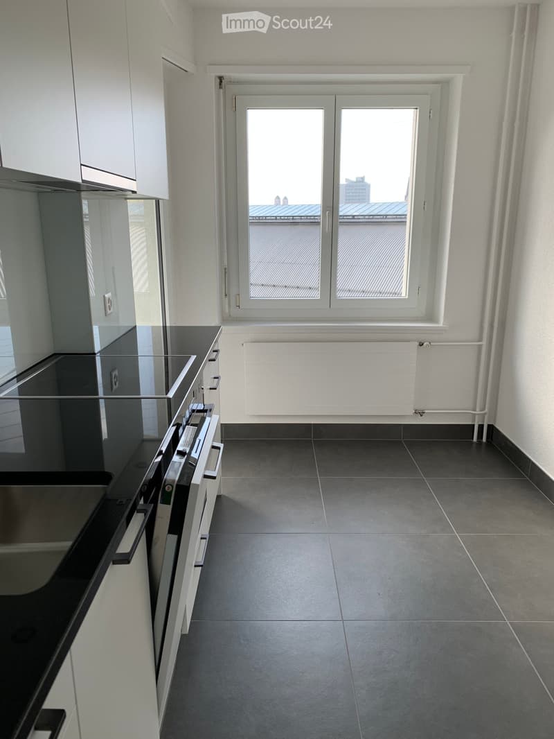 Empty kitchen with dark grey tiled floor, white walls, and a large window overlooking a rooftop. Minimalist design with a black countertop and white cabinets visible on the left side.