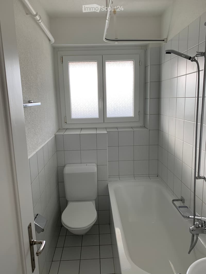 Modern bathroom with white tiled walls and floor, featuring a toilet, bathtub, and a window with frosted glass panes.