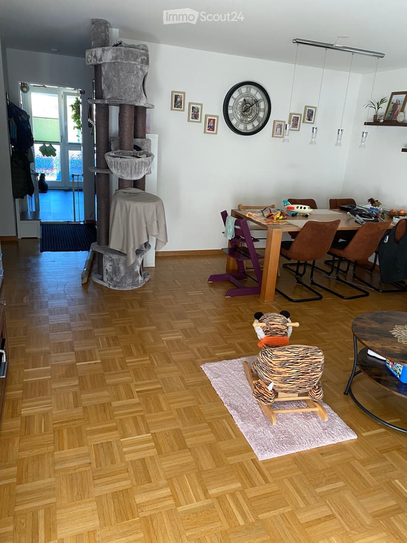 A well-lit room with wooden flooring, a cat tree, a dining table with chairs, a toy on a rug, and a large wall clock.