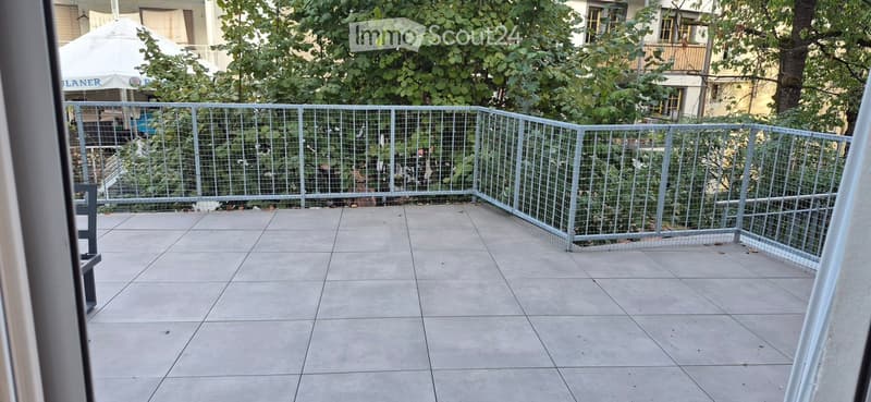 Balcony with tiled floor, metal railing, and greenery visible beyond the railing.