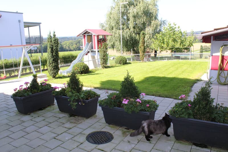 A garden with a brick paved patio, several large potted plants, a slide, and a swing set.