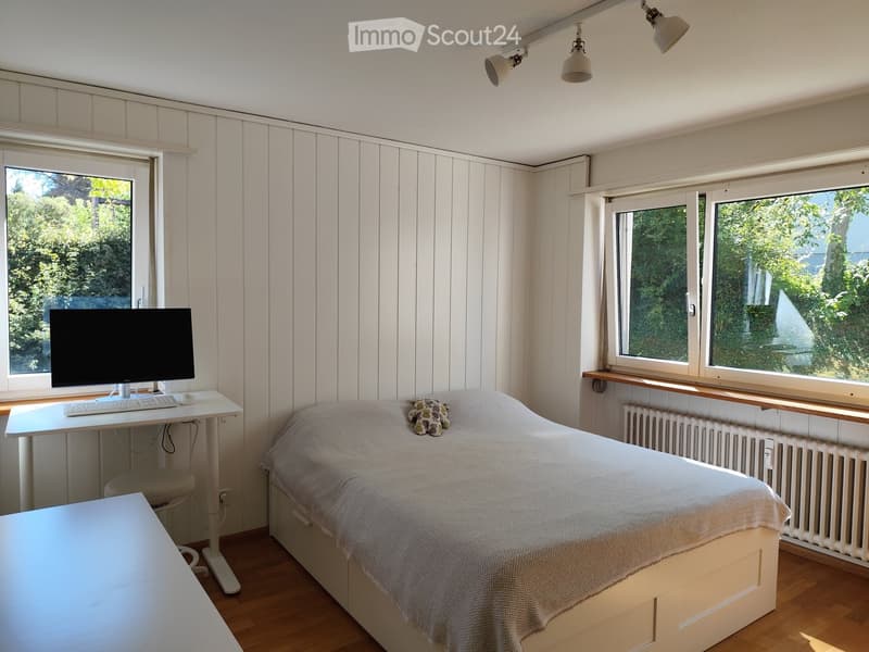 Cozy bedroom with a single bed, a desk with a computer, and a chair. The room features wooden flooring, white paneled walls, and large windows with a view of greenery.