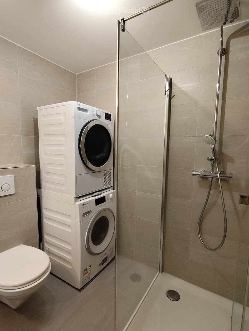 Modern bathroom with a toilet, stacked washing machine, and a glass-enclosed shower with a handheld showerhead.