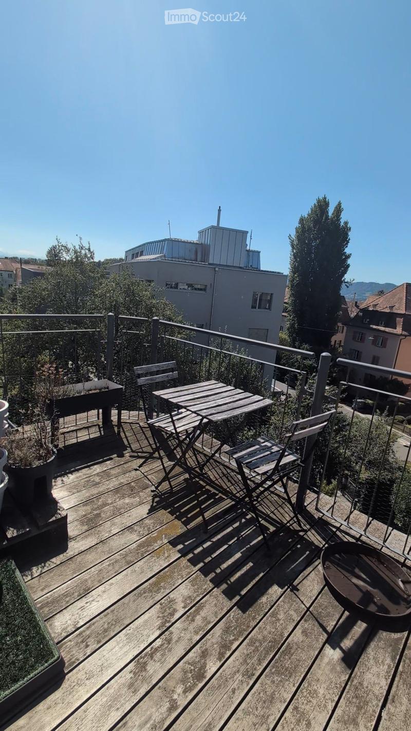 Wooden balcony with a small table, chairs, potted plants, and a fire pit, overlooking a cityscape with buildings and trees.
