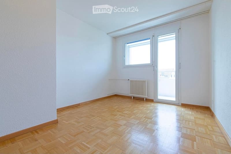 Empty room with wooden parquet flooring, white walls, a radiator, a window, and a glass door leading to a balcony.