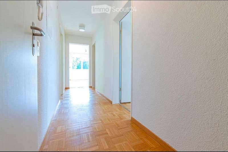 A clean, well-lit hallway with wooden flooring and white walls, featuring a door on the left and a glass door leading to a bright area at the end.