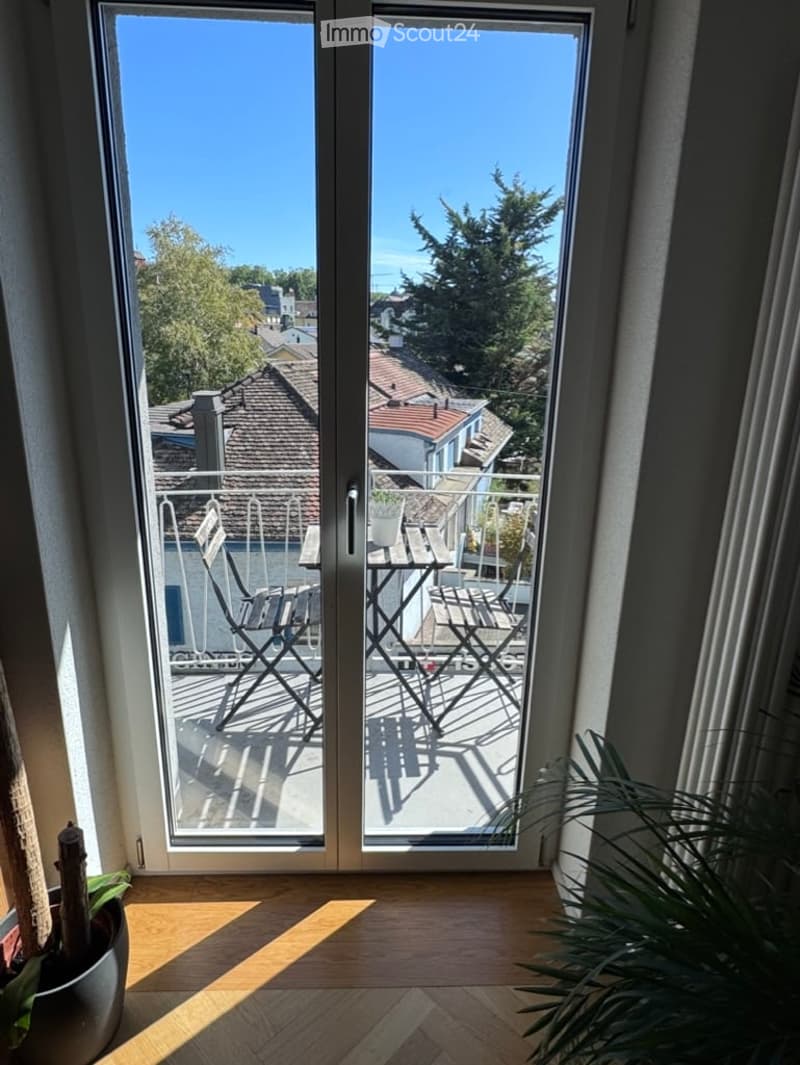 Balcony with outdoor furniture including a table and chairs, overlooking a residential area with trees and rooftops.