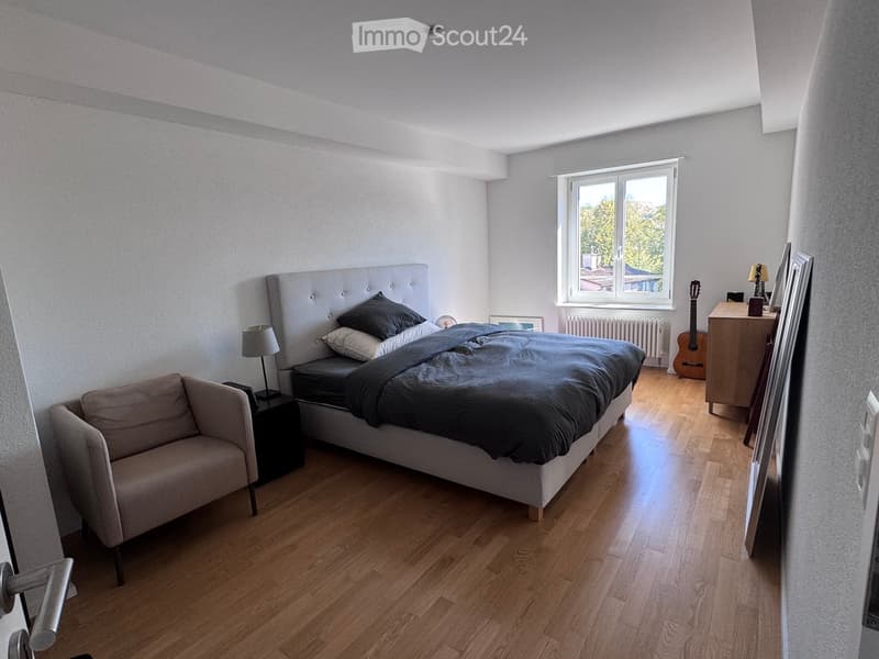 Modern bedroom with wooden flooring, large window, grey upholstered bed, sofa, nightstand, lamp, guitar, and cabinet.