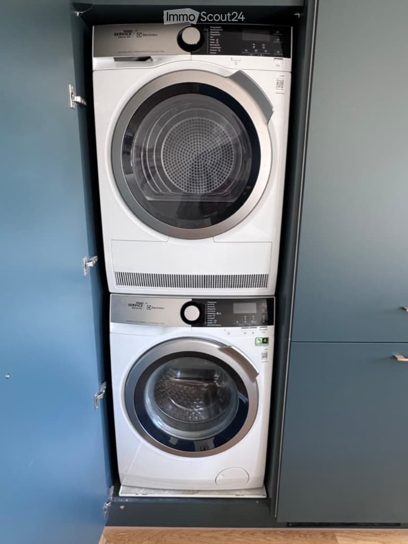 A compact, modern bathroom with a stacked washing machine and dryer unit, featuring sleek white finishes and digital control panels.