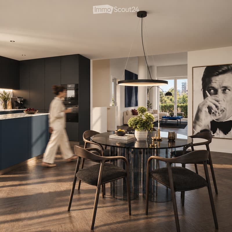 Modern dining area with a round glass table, four chairs, vase with flowers, books, candle, and cup. Adjacent living area features a sofa, coffee table, and plant. Kitchen area visible with dark cabinets, coffee machine, oven, and fridge.