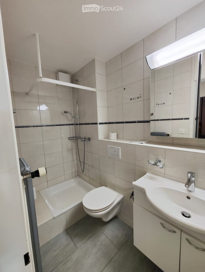 Modern bathroom with a bathtub, toilet, sink, mirror, and showerhead. The walls are tiled in beige with black accents, and the floor is tiled in grey.