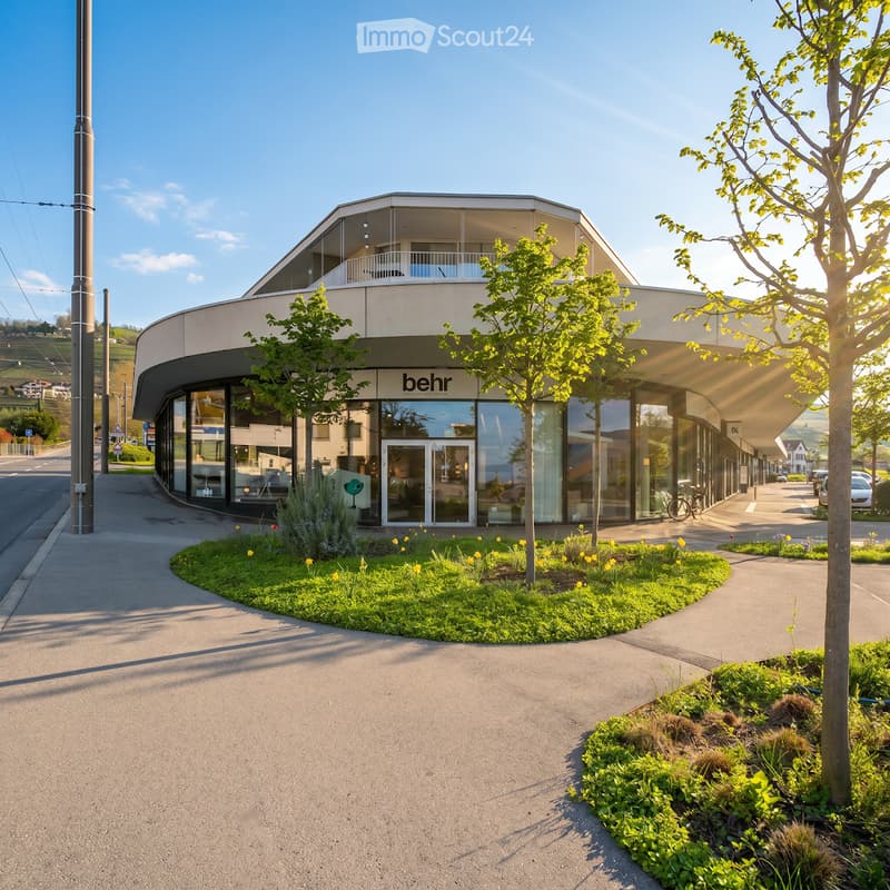 Modern building with a curved facade, large glass windows, and a balcony on the upper level. The entrance features a glass door with the logo "behr" visible. The building is surrounded by greenery, including small trees and flowering plants.