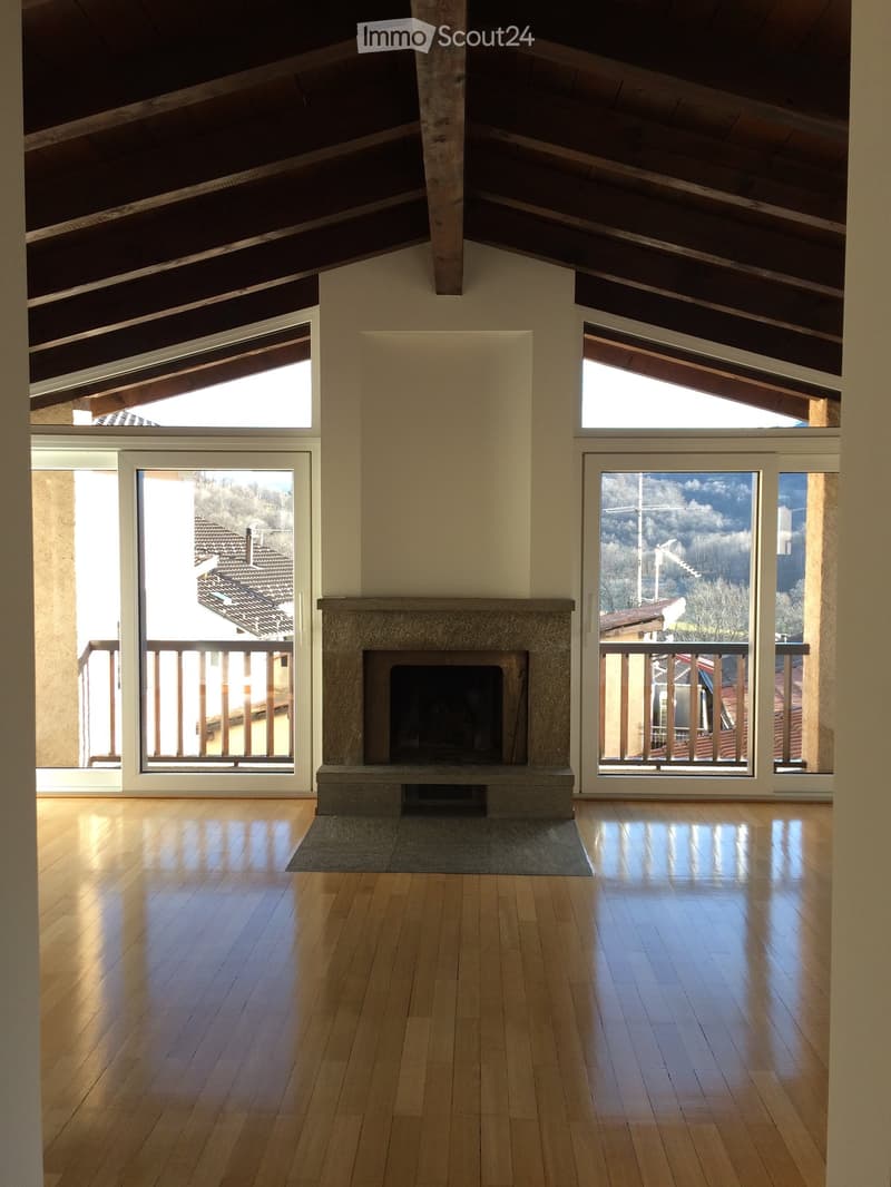 Bright, open living space with wooden floors, large windows, and a central stone fireplace. The room features exposed wooden beams on the ceiling and offers scenic views through the windows.