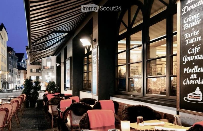 Outdoor seating area of a restaurant with red chairs, tables, and potted plants, illuminated by streetlights.