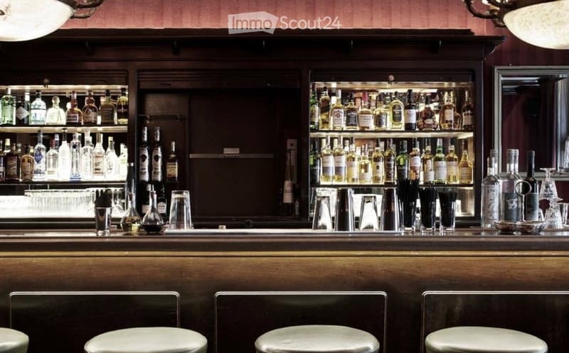 Bar area with wooden counter, shelves filled with bottles, and four white stools.